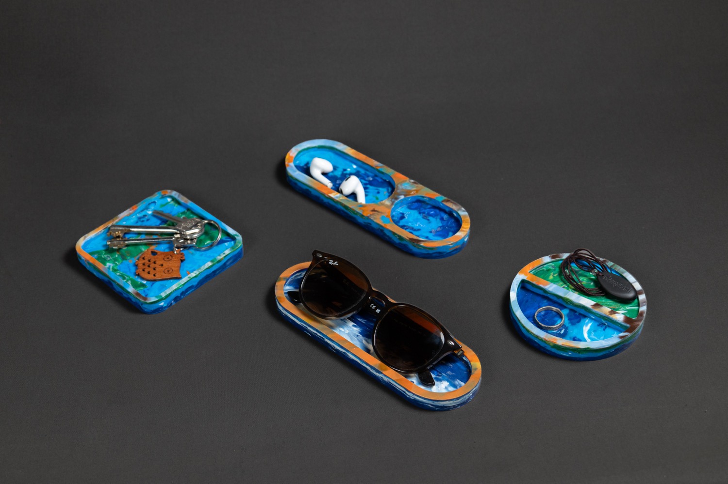Four recycled-plastic trays marbled in blue and orange, holding sunglasses, keys and earrings on a dark grey surface