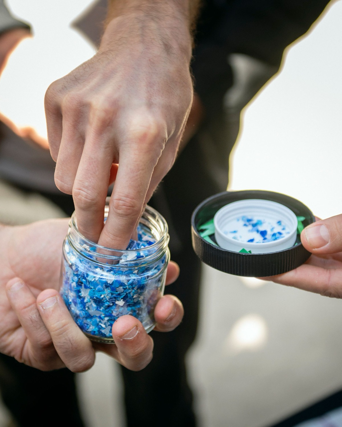 Two people passing a glass jar of blue and white shredded plastic flakes between them
